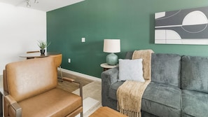 A living room with a green wall, a leather chair, a grey sofa with a throw blanket, a small table with a lamp, and a potted plant. - Landing | Incredible 1BD, Pool (Houston)