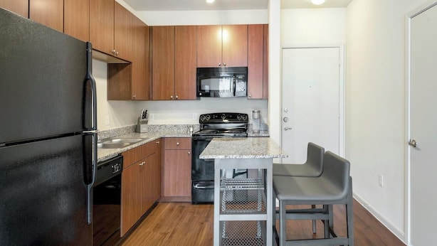 A compact kitchen with wooden cabinets, a stainless steel refrigerator, a microwave, a stove, and a small bar area. - Landing | Amazing 1BD, Pool (Houston)