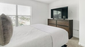 A bedroom with a bed, a dresser, a TV, and a window with blinds. - Landing | Incredible 1BD, Pool (Houston)