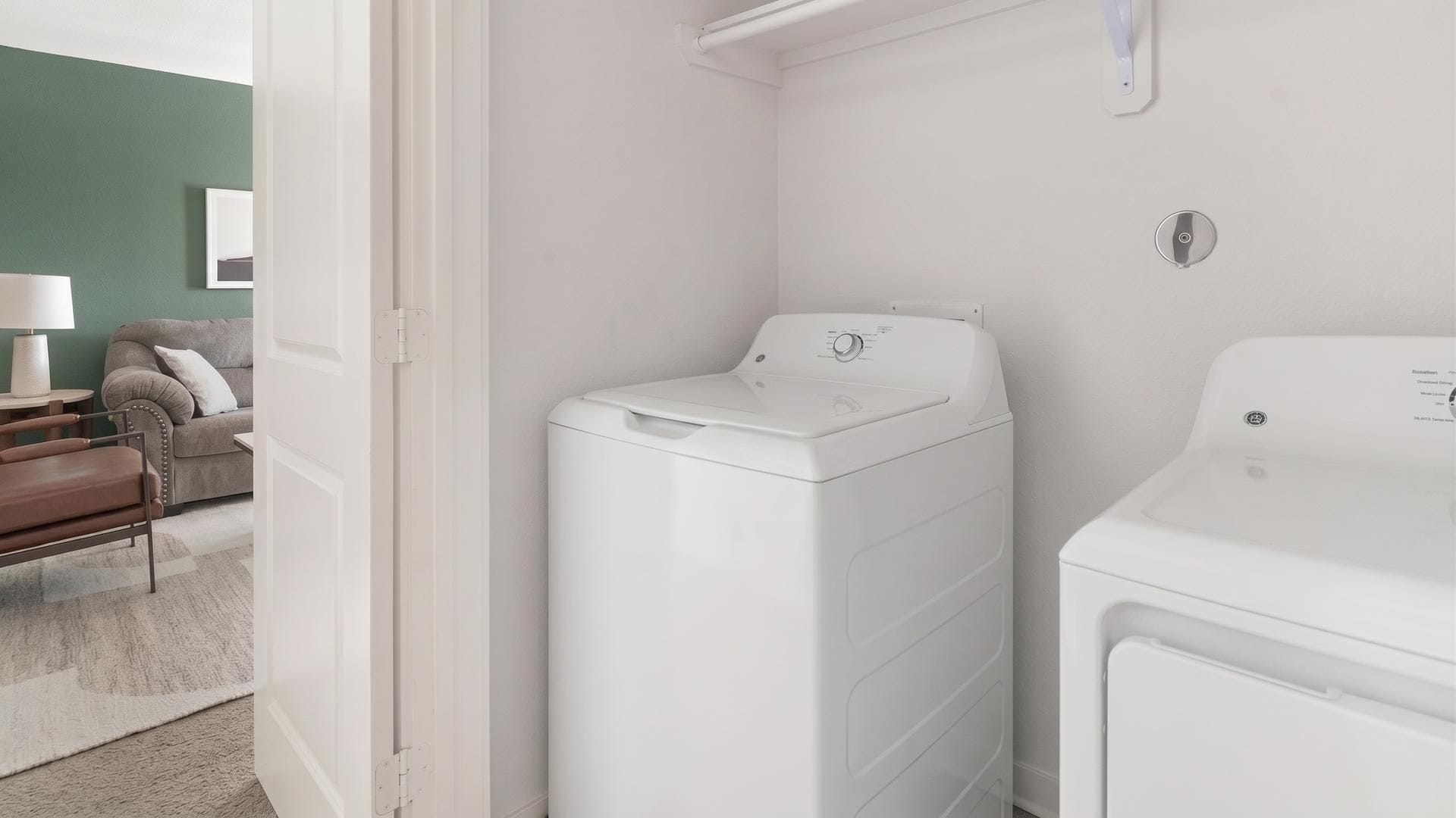 A laundry room with a front-loading washer and dryer, a door leading to a living area with a sofa and a lamp.