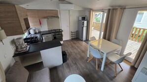 Een compacte keuken met een fornuis, magnetron en koelkast, een eethoek met een tafel en stoelen, en uitzicht op een houten terras.