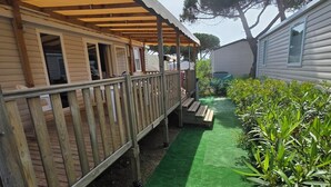 Een overdekt buitengebied met een houten terras, een pad van kunstgras en een omheinde tuin met planten.