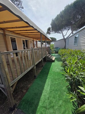 Ein überdachter Außenbereich mit Holzdeck, einem Pfad aus grünem Kunstrasen und einem eingezäunten Garten mit Pflanzen.