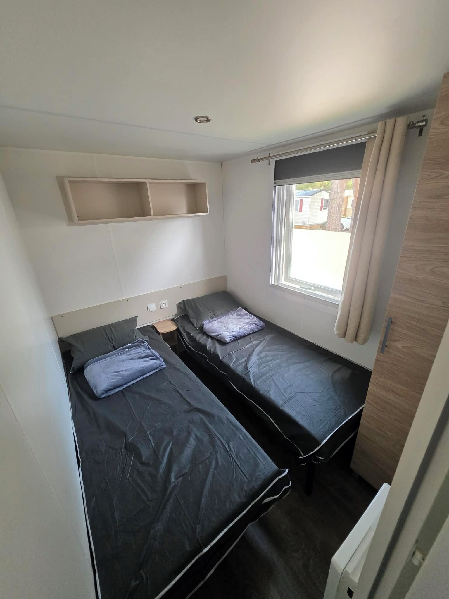 A compact room with two beds, a window with curtains, a shelf above the beds, and a door on the right.