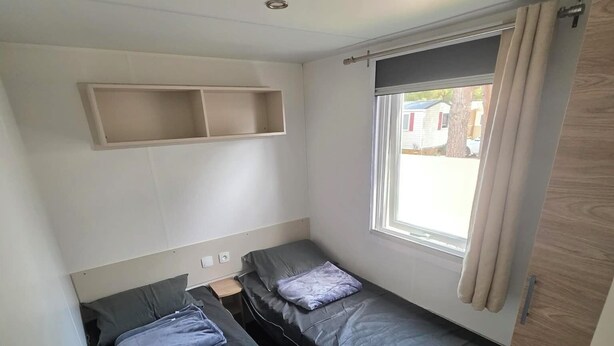 Een kleine kamer met twee bedden, een raam met gordijnen, een plank boven de bedden, en een deur aan de rechterkant.