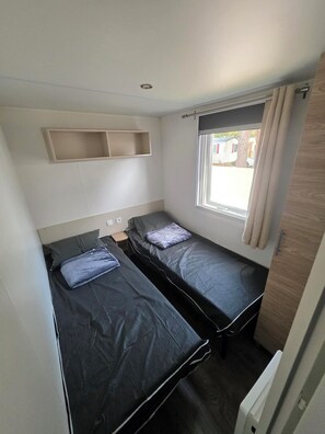A compact room with two beds, a window with curtains, a shelf above the beds, and a door on the right. - Southern Wonder (Canet-en-Roussillon)