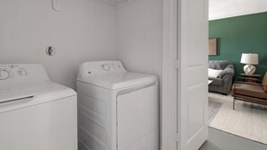 Ein Waschraum mit zwei weißen Frontlader-Waschmaschinen und Trocknern, eine Tür zu einem Wohnbereich mit einer Couch und einer Lampe.