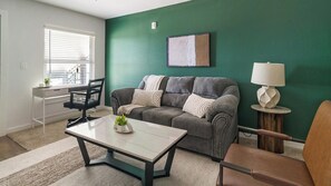 A living room with a green wall, a grey sofa, a wooden side table, a white lamp, and a small plant on a coffee table. - Landing | Chic 1BD, Pool (Houston)