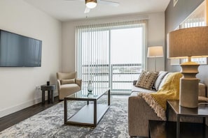A modern living room with a glass coffee table, a beige sofa, a television mounted on the wall, and a large window with blinds. - Landing | Incredible 1BD, Pool (Houston)