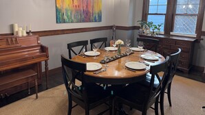 Comedor con una mesa de madera dispuesta para cuatro, sillas, un piano y un cuadro abstracto grande en la pared.