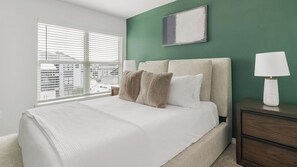 Une chambre à coucher avec un lit, des oreillers, une table de chevet, une lampe et une vue sur les bâtiments par la fenêtre.