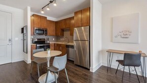 A modern kitchen with stainless steel appliances, wooden cabinets, a dining area with a round table and chairs, and a wall-mounted light fixture. - Landing | Stunning 2BD, Pool (Houston)