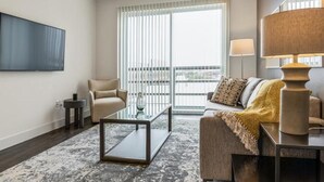 A modern living room with a glass coffee table, a beige sofa, a television mounted on the wall, and a large window with blinds. - Landing | Modern 1BD, Pool (Houston)