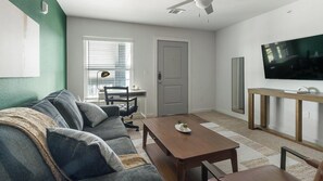 A living room with a sofa, a coffee table, a TV, and a door. - Landing | Chic 1BD, Pool (Houston)