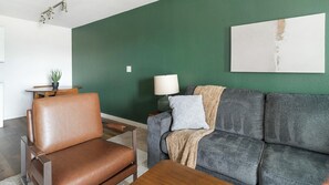A living room with a brown leather chair, a grey sofa with a blanket, a small table, a lamp, and a green wall. - Landing | Chic 1BD, Pool (Houston)