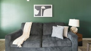 A grey sofa with a white throw blanket and a patterned pillow against a green wall. A framed abstract artwork is above the sofa, and a white lamp is on a wooden side table beside it. - Landing | Amazing 1BD, Pool (Houston)