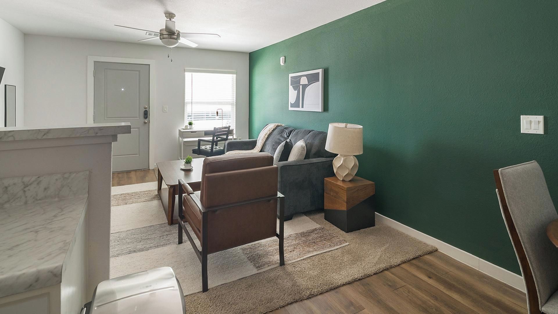A modern living room with a green accent wall, a ceiling fan, a dining area with a table and chairs, and a view of the outdoors.