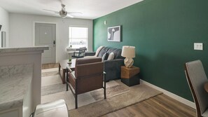 Un salon moderne avec un mur d’accent vert, un ventilateur de plafond, un coin repas avec une table et des chaises, et une vue sur l’extérieur.
