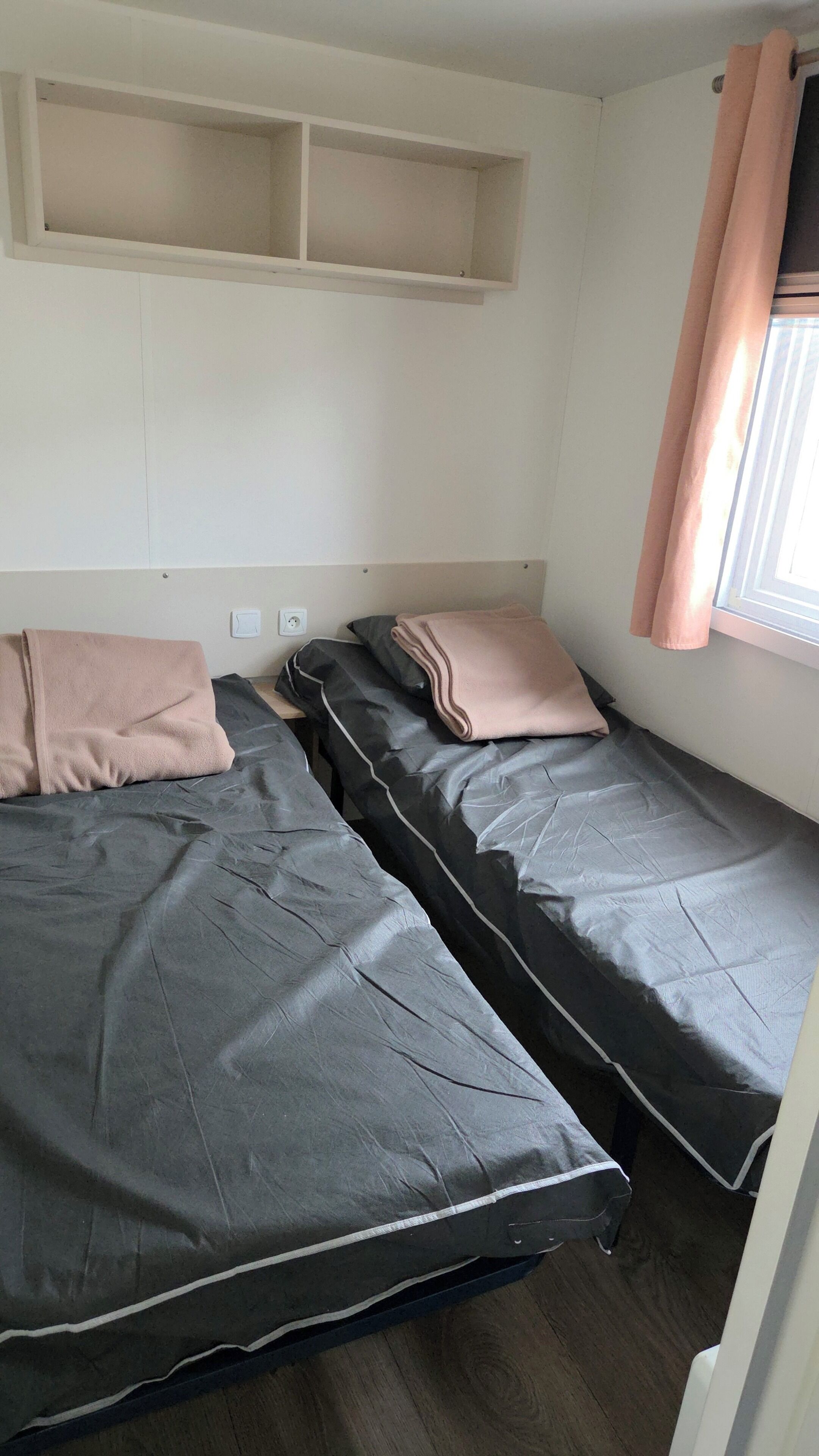 A bedroom with two single beds, a window with curtains, and a shelf above the beds.