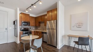 A modern kitchen with wooden cabinets, stainless steel appliances, a dining area with a round table and chairs, and a wall-mounted light fixture. - Landing | Stylish 2BD, Pool (Houston)