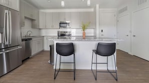 A modern kitchen with stainless steel appliances, a marble countertop, and two black bar stools. - Landing | Spectacular 1BD, Gym, Yoga Studio (Sarasota)