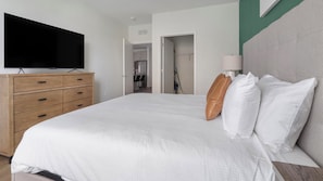 A hotel room with a large bed, a flat-screen TV mounted on a wooden dresser, and a door leading to a bathroom. - Landing | Chic 1BD, Yoga Studio, Gym (Sarasota)