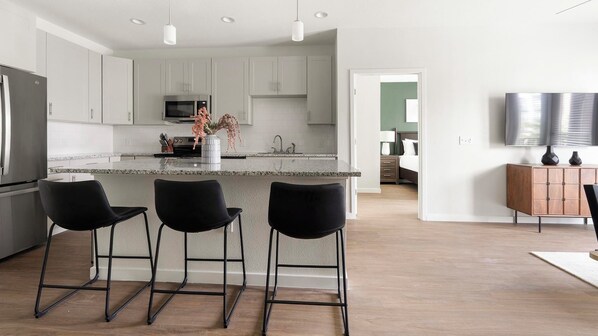 A modern kitchen with a central island, black bar stools, and a dining area with a wooden cabinet and a flat-screen TV. - Landing | Spectacular 2BD, Yoga Studio, Gym (Sarasota)