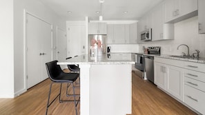 A modern kitchen with white cabinets, a stainless steel refrigerator, a microwave, and a sink. There are two black bar stools and a vase with flowers on the island. - Landing | Spectacular 2BD, Yoga Studio, Gym (Sarasota)