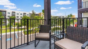 A balcony with two wicker chairs and a table, overlooking a parking area and buildings. - Landing | Spectacular 2BD, Yoga Studio, Gym (Sarasota)
