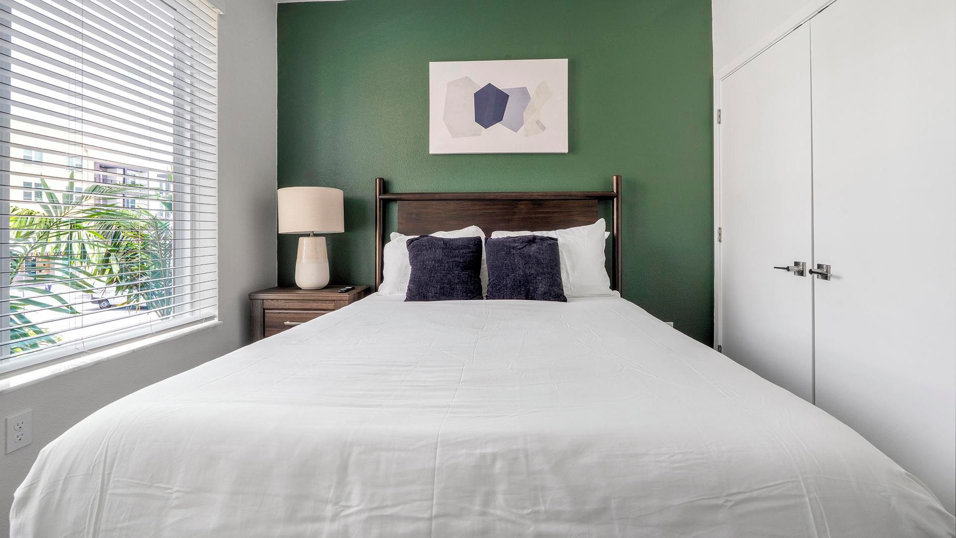 Une chambre d’hôtel avec un lit, une table de chevet avec une lampe, un mur vert et une fenêtre avec des stores.