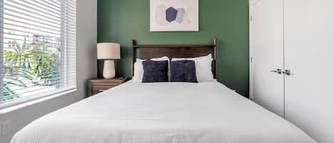 Une chambre d’hôtel avec un lit, une table de chevet avec une lampe, un mur vert et une fenêtre avec des stores.