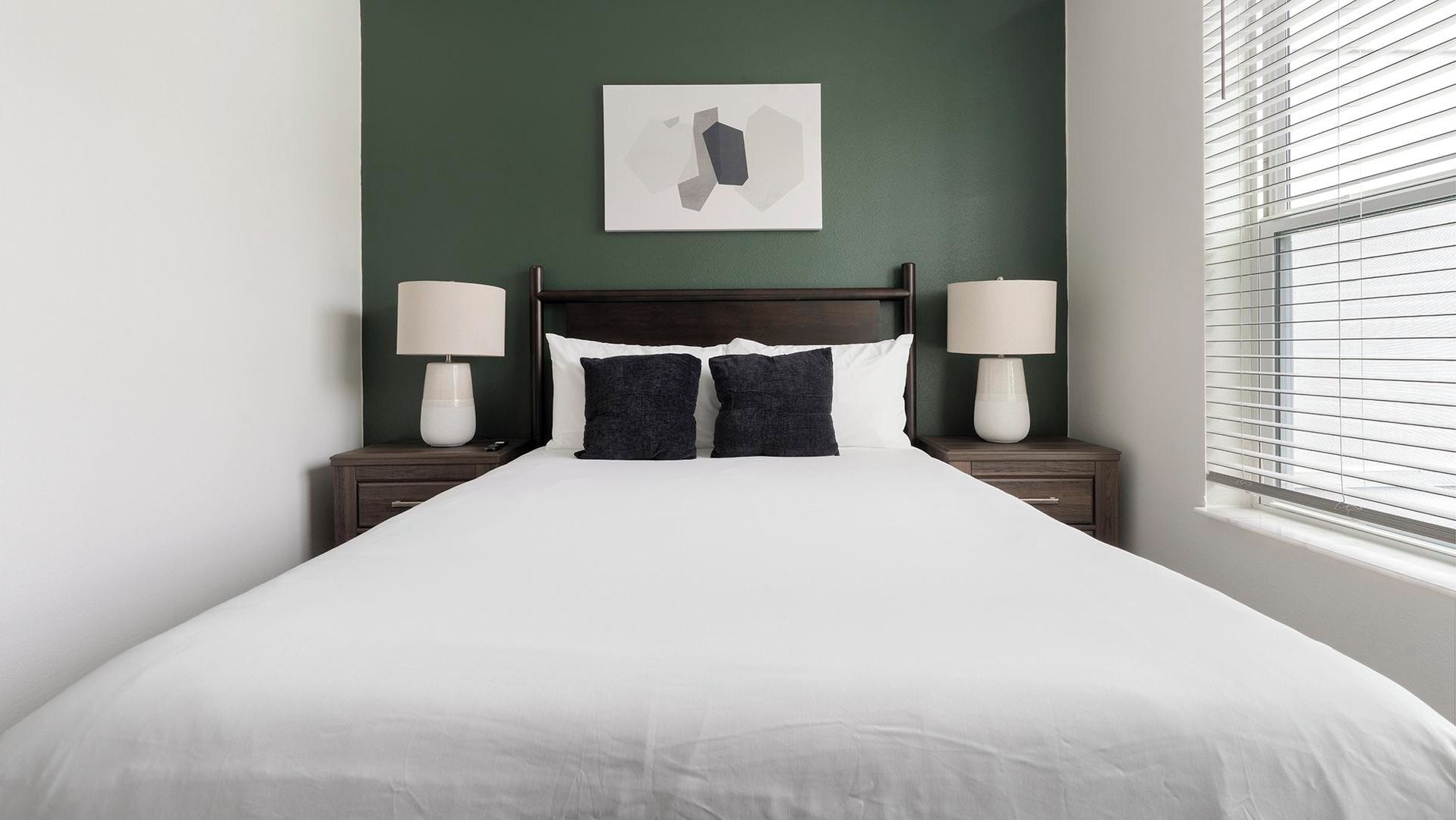 Quarto de hotel com uma cama grande, duas mesas de cabeceira com abajures, uma parede verde escura e uma janela com persianas.