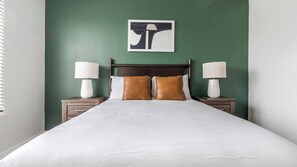 A neatly made bed with white bedding and two brown pillows, flanked by nightstands with lamps, against a green wall with a framed artwork above. - Landing | Chic 1BD, Yoga Studio, Gym (Sarasota)