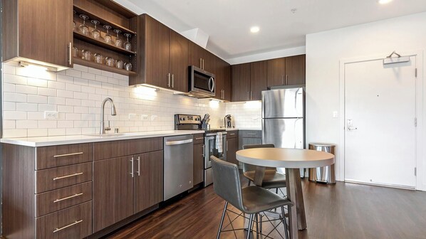 A modern kitchen with dark wood cabinets, stainless steel appliances, a white tiled backsplash, and a round dining table with two chairs. - Landing | Incredible 1BD, Yoga Studio, Gym (Sarasota)
