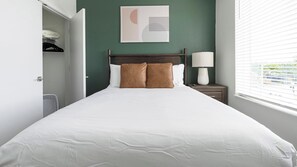 Quarto de hotel com cama, mesa de cabeceira com luminária, obra de arte na parede e janela com persianas.