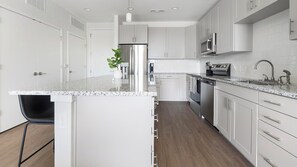A modern kitchen with white cabinetry, a granite countertop, stainless steel appliances, and a black chair. - Landing | Beautiful 2BD, Gym, Yoga Studio (Sarasota)