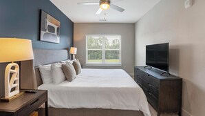 Un dormitorio con cama, televisor, ventilador de techo y ventana con persianas.