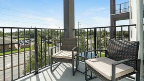 Een balkon met twee rieten stoelen en een kleine tafel, met uitzicht op een straat met geparkeerde auto's en gebouwen.