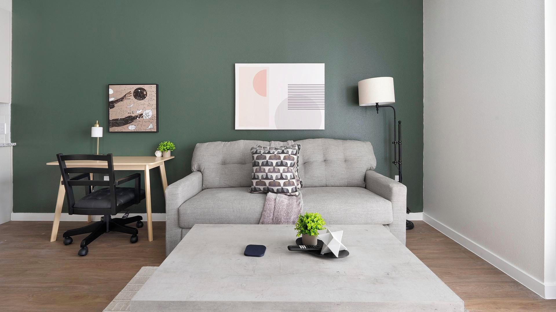 A living room with a grey sofa, a small table, a desk with a chair, a lamp, and a framed abstract artwork on the wall.