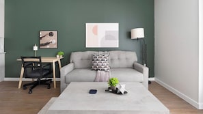 Ein Wohnzimmer mit grauem Sofa, einem kleinen Tisch, einem Schreibtisch mit Stuhl, einer Lampe und einem gerahmten abstrakten Kunstwerk an der Wand.