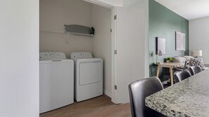 A laundry room with two front-loading washers and dryers, a small shelf above the dryers, and a dining area with a table and chairs. - Landing | Amazing 2BD, Gym, Yoga Studio (Sarasota)