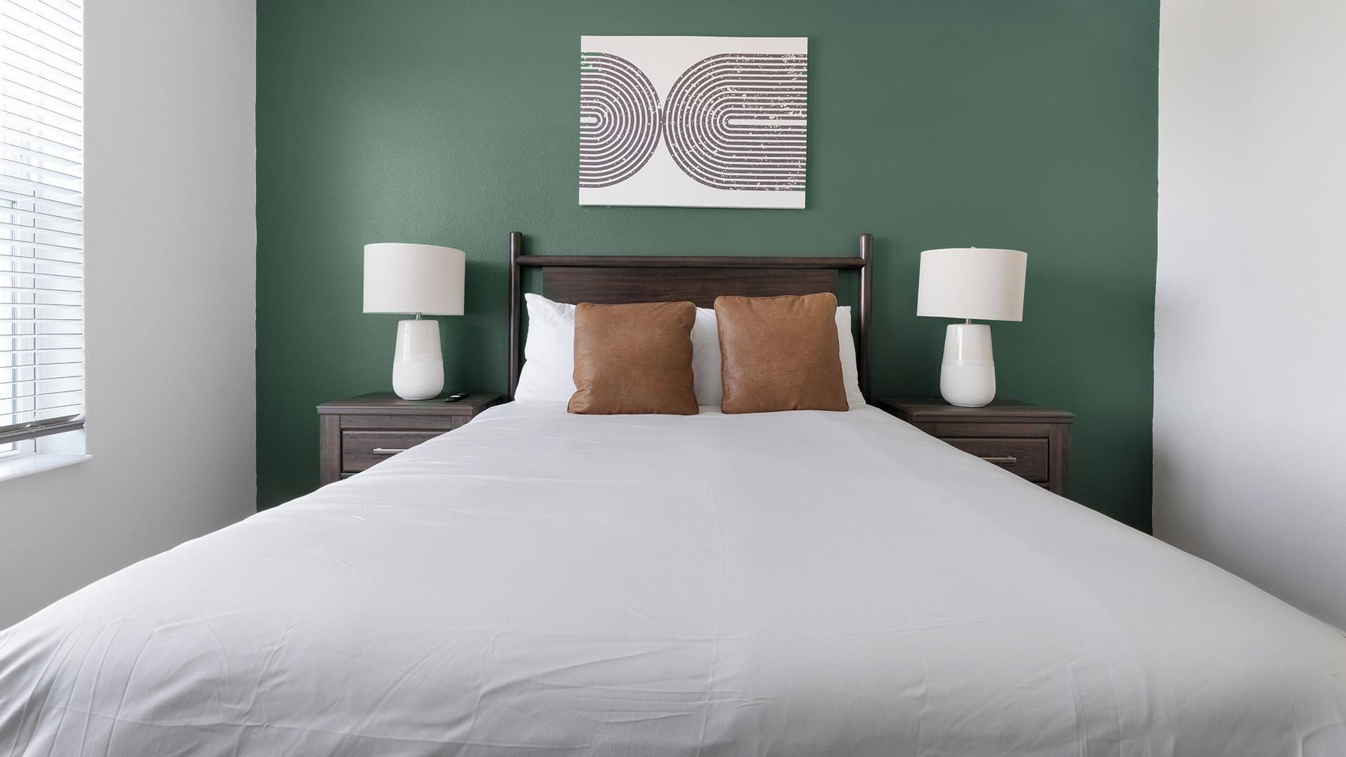 Une chambre d’hôtel avec un grand lit, deux tables de chevet avec des lampes, un mur d’accent vert et une œuvre d’art abstraite au-dessus du lit.