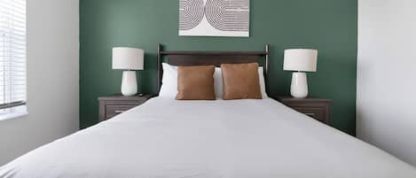 Une chambre d’hôtel avec un grand lit, deux tables de chevet avec des lampes, un mur d’accent vert et une œuvre d’art abstraite au-dessus du lit.