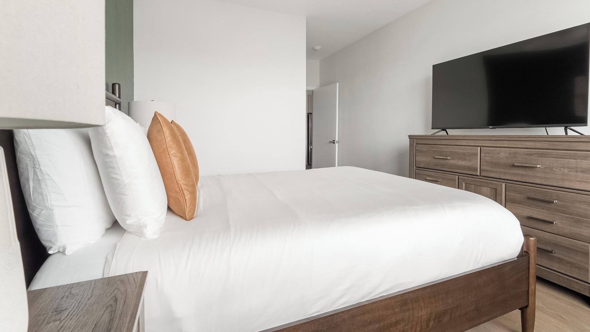A modern bedroom with a bed, a television on a wooden dresser, and a door in the background.