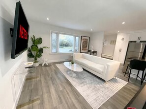 A modern living room with a white sofa, a coffee table, and a TV mounted on the wall. - 5BR · Sleeps 10 · Parking · Near Rogers Stadium (Toronto)