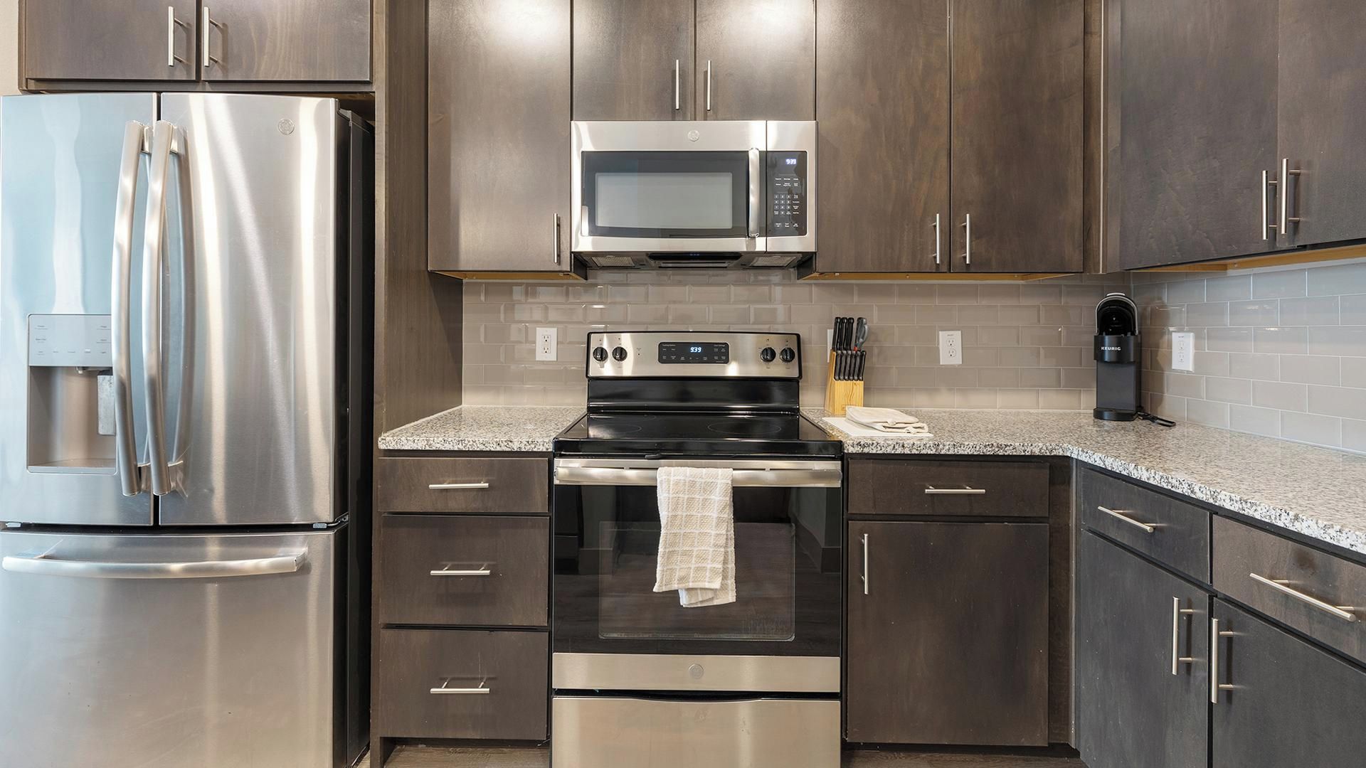 Una cocina con electrodomésticos de acero inoxidable, que incluyen un refrigerador, microondas y horno, con gabinetes de madera oscura y encimera de granito.
