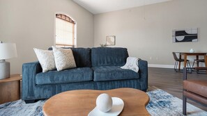 A living room with a sofa, a coffee table, a dining table, and a window with blinds. - Landing | Stylish 1BD, Gym, Courtyard (San Antonio)