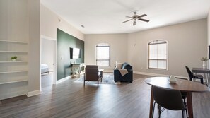 A spacious living room with a ceiling fan, a dining area with a round table, and a bedroom with a bed and a TV. - Landing | Incredible 1BD, Courtyard, Gym (San Antonio)