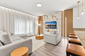 Een moderne woonkamer met een zithoek, een salontafel, een aan de muur bevestigde televisie en een eethoek met krukken.