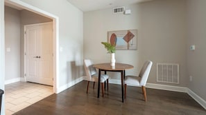 A dining area with a round table, two chairs, a vase with a plant, and a wall-mounted abstract painting. - Landing | Stylish 1BD, Courtyard, Gym (San Antonio)
